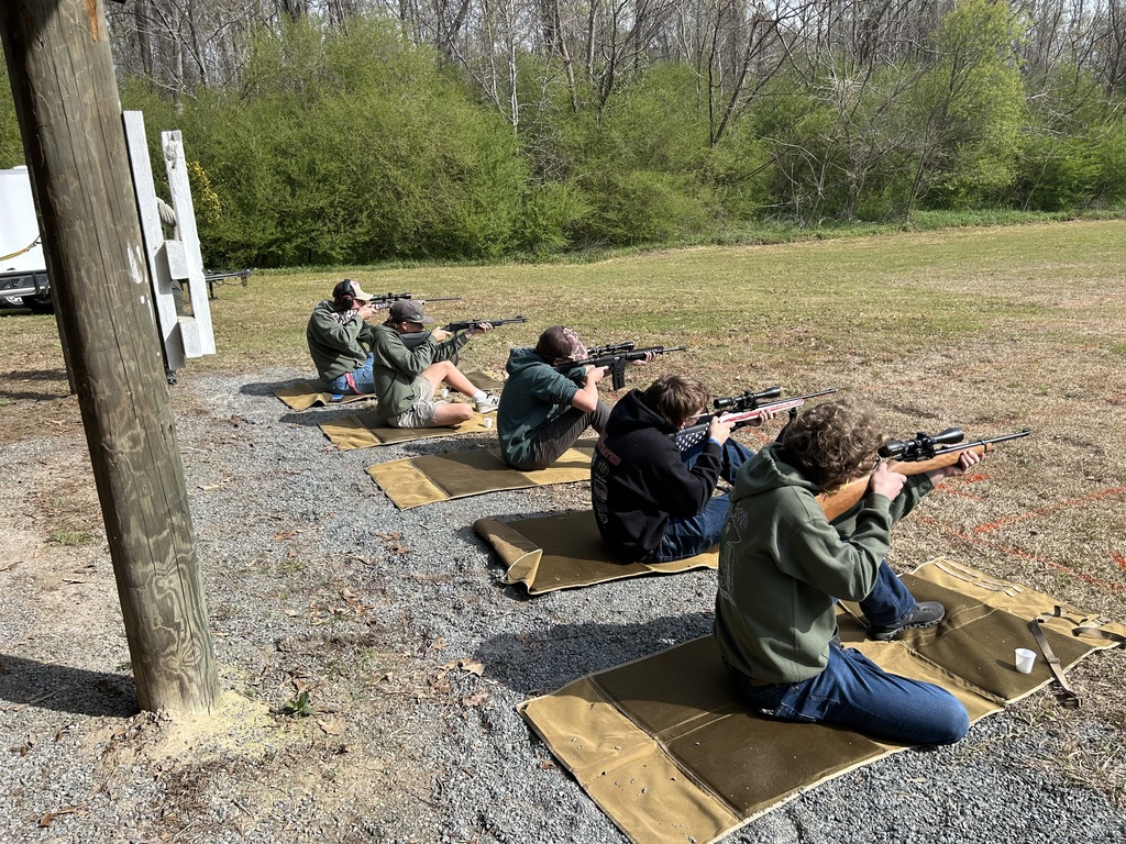 Four students wearing casual outdoor clothing and safety ear protection participate in a target shooting competition. They are kneeling on brown mats in a grassy field, aiming rifles toward a treeline under a clear sky