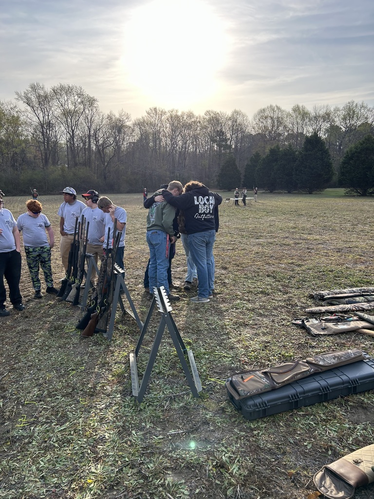 A group of high school students and instructors standing in a grassy field during an outdoor target practice session. Several students are positioned at wooden shooting benches with equipment, while others stand behind them in conversation. The background features a dense line of autumn trees under a hazy, overcast sky