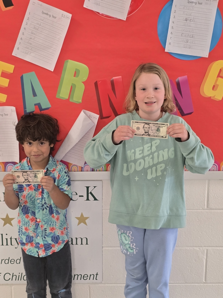 Two elementary-aged students stand in front of a bright red bulletin board decorated with colorful letters spelling “EARNING” and posted spelling tests. Each student is smiling and holding up a five-dollar bill. One student wears a green sweatshirt and blue pants, while the other wears a colorful patterned shirt and dark pants. A sign partially visible behind them references a Pre-K program and student achievement.