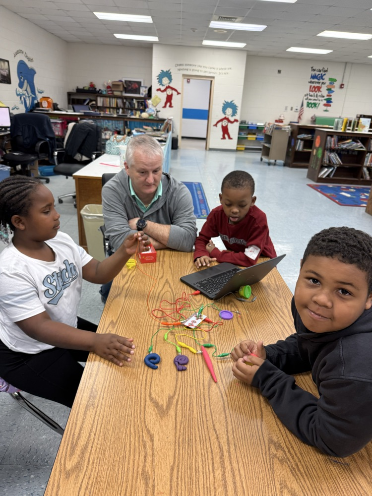 Students and an adult sit around a table in a classroom working on a hands-on STEM activity using wires, clay, and a laptop, collaborating and learning together.