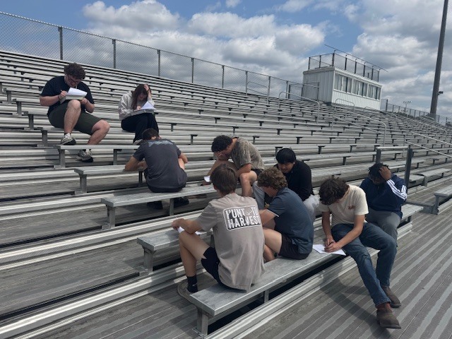 A group of high school students sitting on outdoor bleachers, focused on writing in notebooks during an outdoor class session