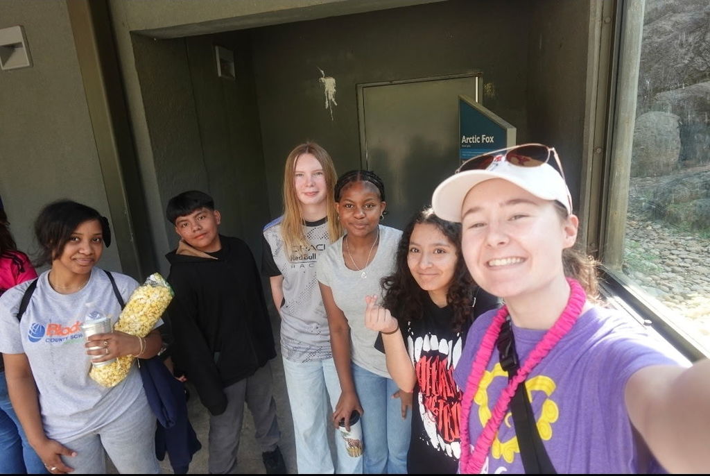 A group of students takes a selfie near a zoo exhibit entrance, smiling and holding snacks and drinks.