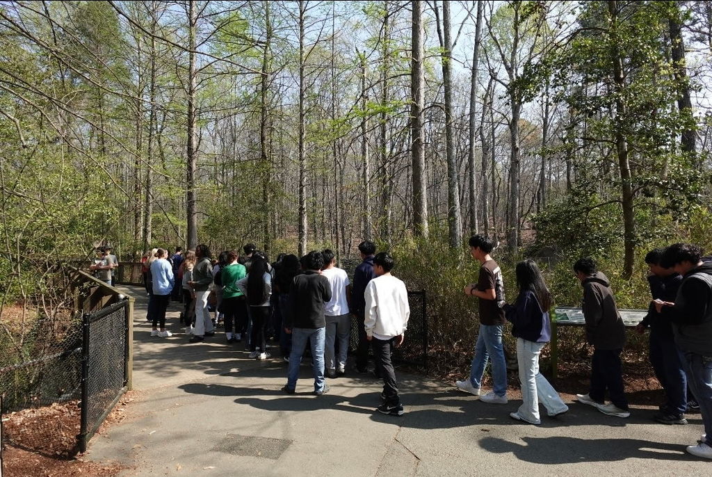 A line of students walks along a wooded path at a zoo, following a guide toward an exhibit.