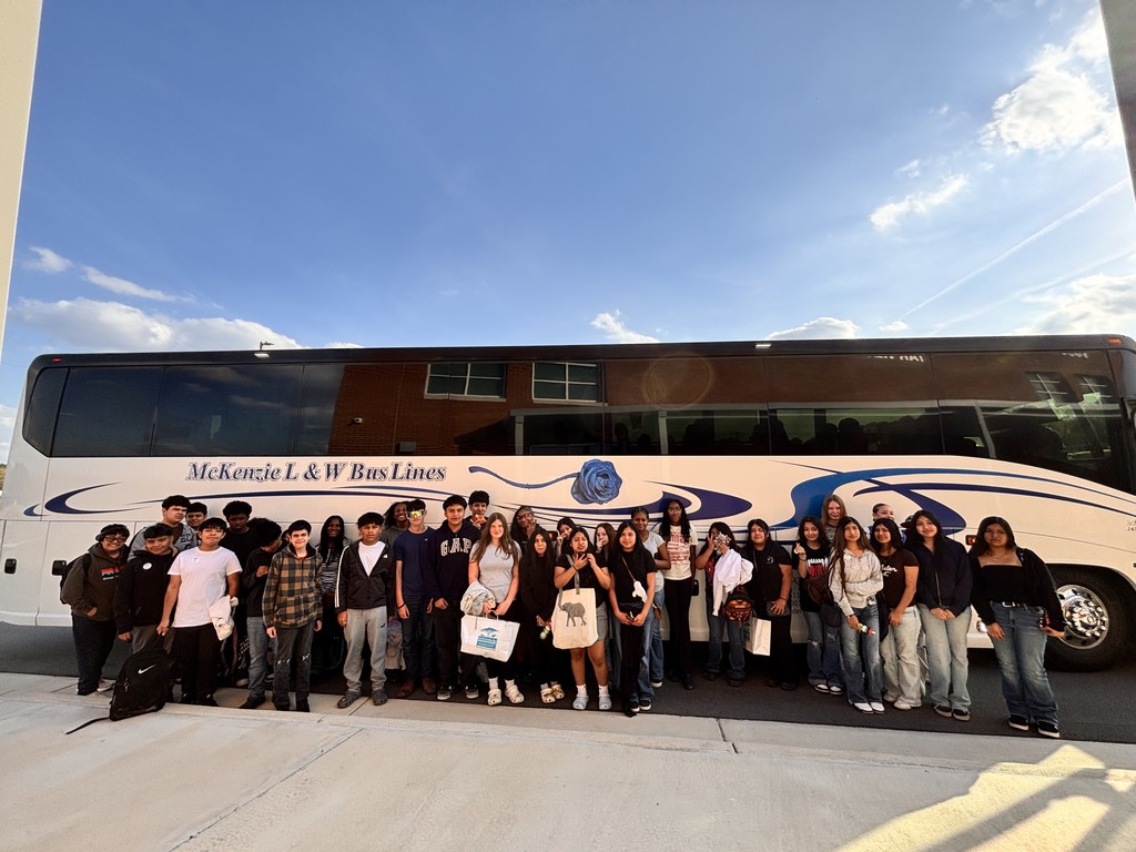 A large group of students stands together in front of a white charter bus outside a school building on a sunny day.