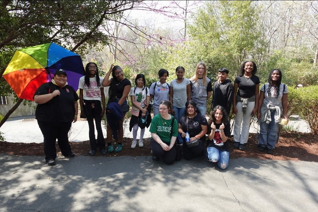 A group of students poses together outdoors on a paved path surrounded by trees, with one person holding a colorful umbrella.