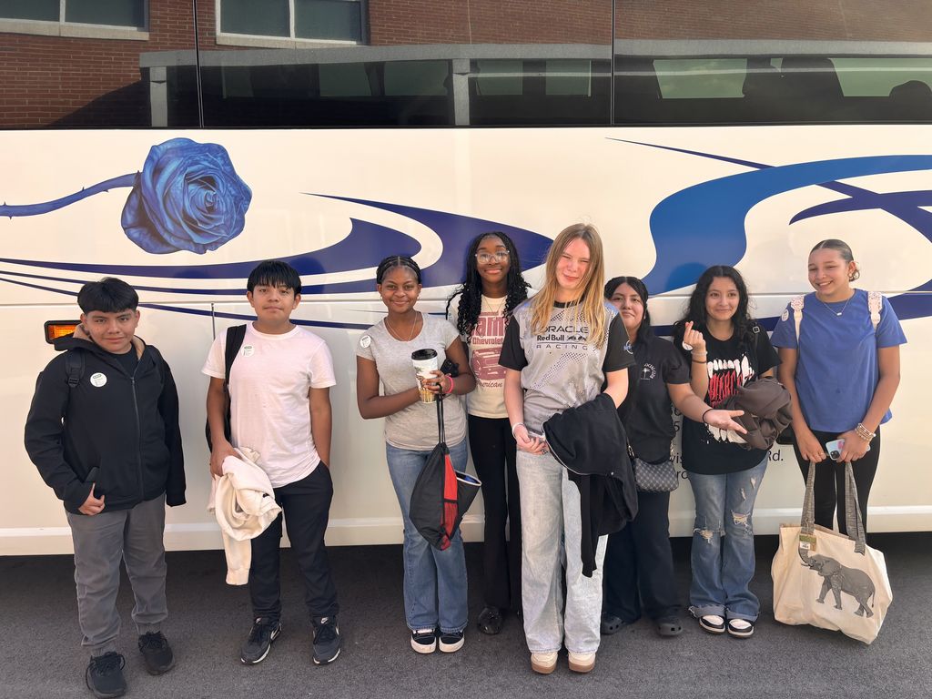 A smaller group of students poses and smiles in front of a charter bus, holding bags and personal items.