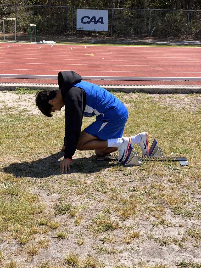 Male student in blue track uniform knealing on the ground to stretch and prepare to run at a track meet.