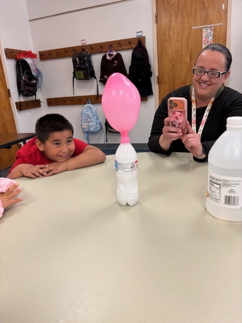 Students watch as a balloon inflates on top of a bottle during a classroom experiment demonstrating a chemical reaction that produces gas.