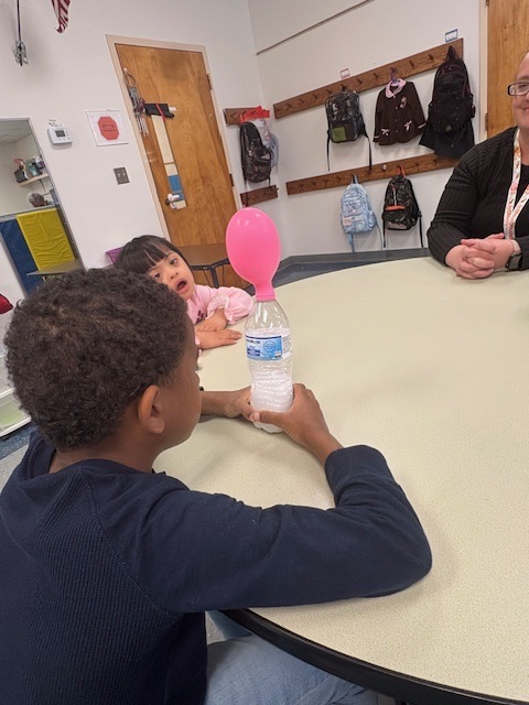 Students watch as a balloon inflates on top of a bottle during a classroom experiment demonstrating a chemical reaction that produces gas.
