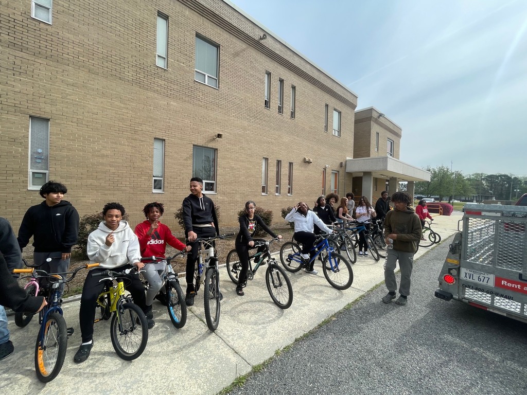 A group of teenagers line up with bicycles along a sidewalk beside a brick school building, some sitting on their bikes and others standing, with a U-Haul trailer parked nearby.