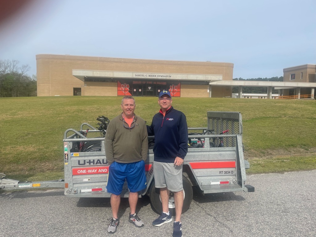 Two men stand side by side in front of a U-Haul trailer loaded with bicycles, parked on a paved area in front of a large school gymnasium building. The building behind them has a wide entrance with red accents and signage, and sits on a grassy hill under a clear blue sky.