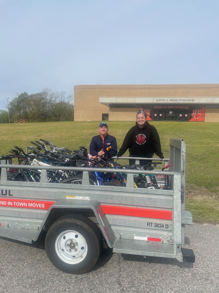 Two adults stand behind a U-Haul trailer filled with bicycles, smiling at the camera in front of a school gymnasium building on a grassy campus under a clear sky.