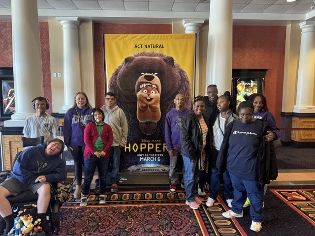 Students are standing in front of the Hoppers movie poster at Millstone Theatre in Hope Mills.From left to right, young man sitting in his wheelchair. behind him is a youg man wearing purple headphones. A young lady standing beside him wearing a purple sweatshirt and another in a red sweater. On the other side of the sign is a young man wearing a West Bladen Knights hoodie, a young man wearing a black hoodie and a parent volunteer wearing a black jacket.