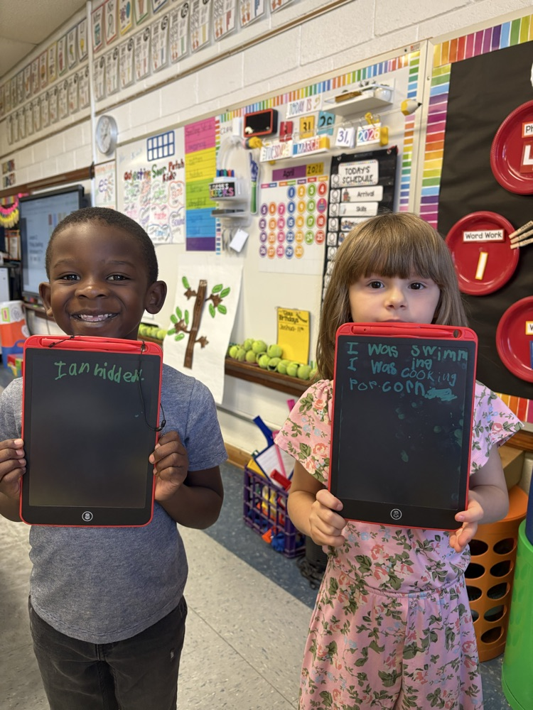 Two students smile while holding writing tablets showing their sentences in a classroom.