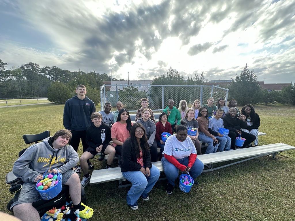 A group of approximately 20 students and staff members sitting on outdoor bleachers and standing together for a group photo. Many students are holding colorful Easter baskets. They are gathered in front of a chain-link fence with a school building and a cloudy sky in the background."