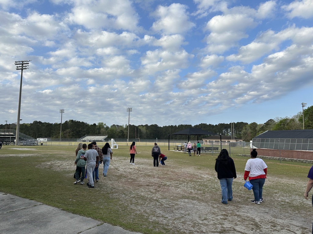 A group of students and staff gathered on a sandy field at West Bladen High School under a bright blue sky with wispy white clouds. In the background, a brick school building, stadium lights, and a black canopy tent are visible