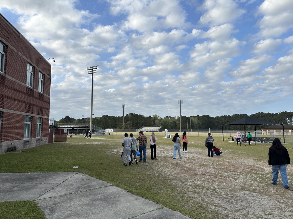 A group of students and staff gathered on a sandy field at West Bladen High School under a bright blue sky with wispy white clouds. In the background, a brick school building, stadium lights, and a black canopy tent are visible.