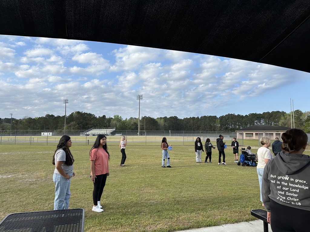 A grassy field with several colorful Easter eggs scattered in the foreground. In the background, a group of people is gathered around black metal picnic tables under a large black square sunshade canopy. A low-profile brick building and a line of trees are visible under a cloudy sky.