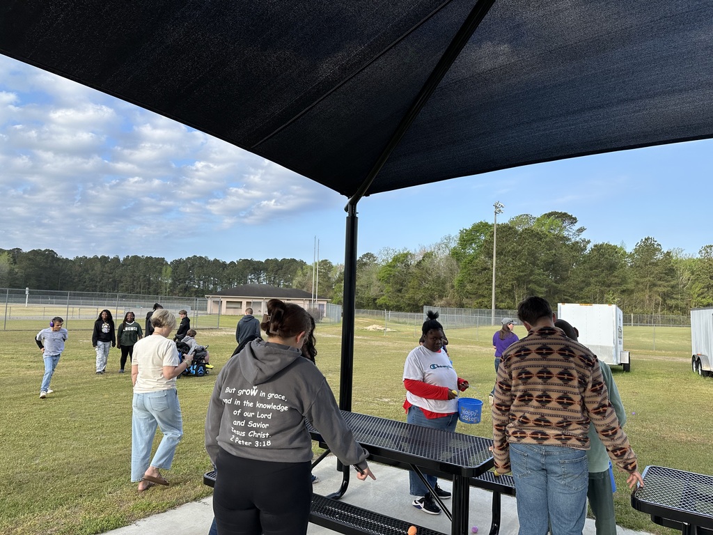 A group of students and staff gathered under a large black shade canopy on a grassy field at West Bladen High School. Students are interacting near a blue table, with athletic fields and trees visible in the background under a clear sky