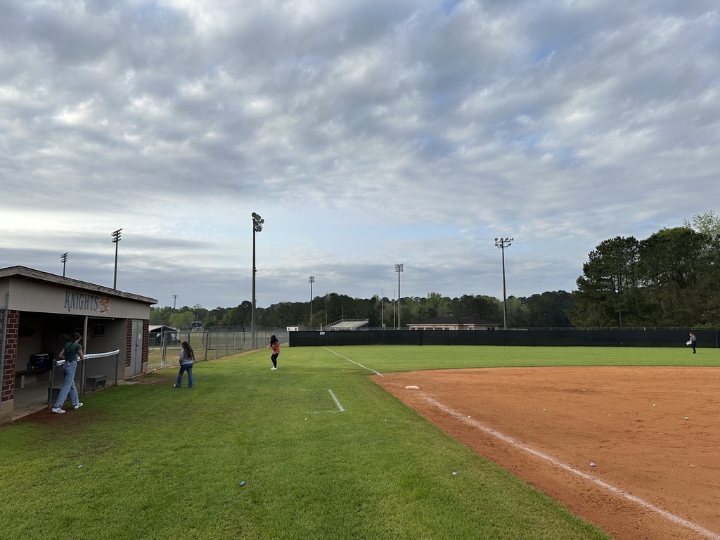 A view of the West Bladen High School baseball field under a cloudy sky. Three students are walking near the dugout, which is labeled with 'Knights' in purple and gold lettering