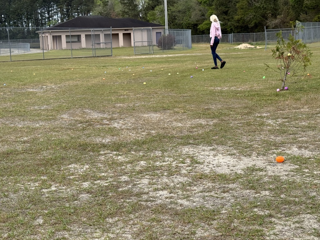 A wide outdoor shot of an Easter egg hunt on a grassy field. A person in a pink shirt and dark pants walks across the field, which is scattered with colorful plastic eggs. In the background, there is a brick school building, a chain-link fence, and several trees under a lightly clouded sky
