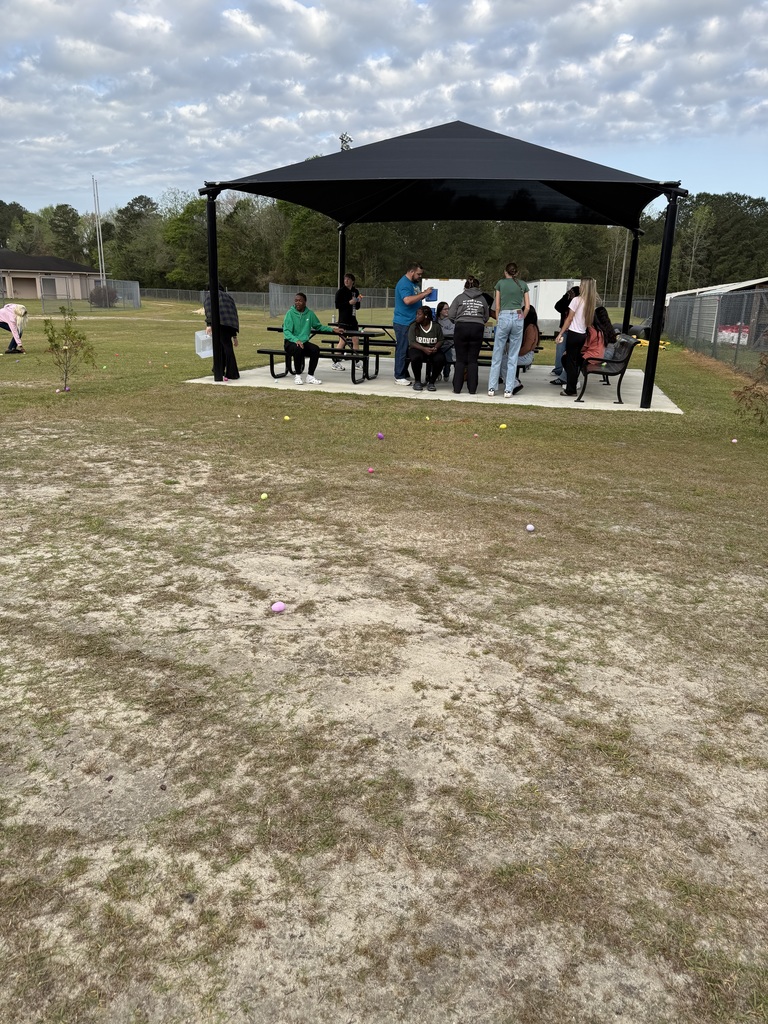 A group of people gathering around black metal picnic tables under a large black square sunshade canopy on a grassy field. Several individuals are standing and talking while others sit. In the background, there are trees and a low-profile brick building under a cloudy sky.