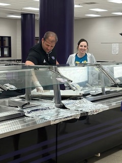 Principal Mr. Orrock, wearing a black polo shirt and gloves, serves food behind a school cafeteria counter while a female cafeteria worker in a hairnet and blue shirt smiles alongside him