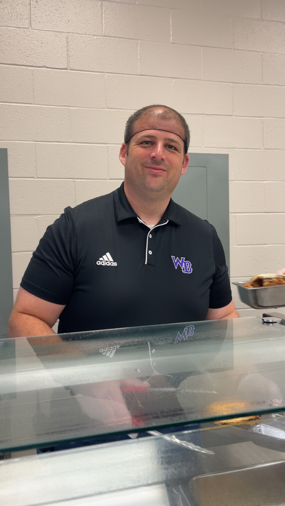 A smiling male staff member wearing a black polo shirt and a hairnet, leaning his head on his hand while standing behind a school cafeteria serving counter.