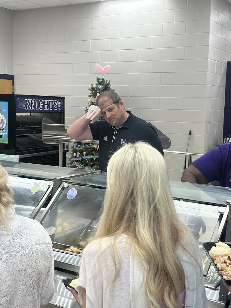 A male staff member in a black polo shirt smiling and leaning his head on his hand while standing behind a school cafeteria counter. In the background, a small decorated Christmas tree with pink bunny ears on top is visible.