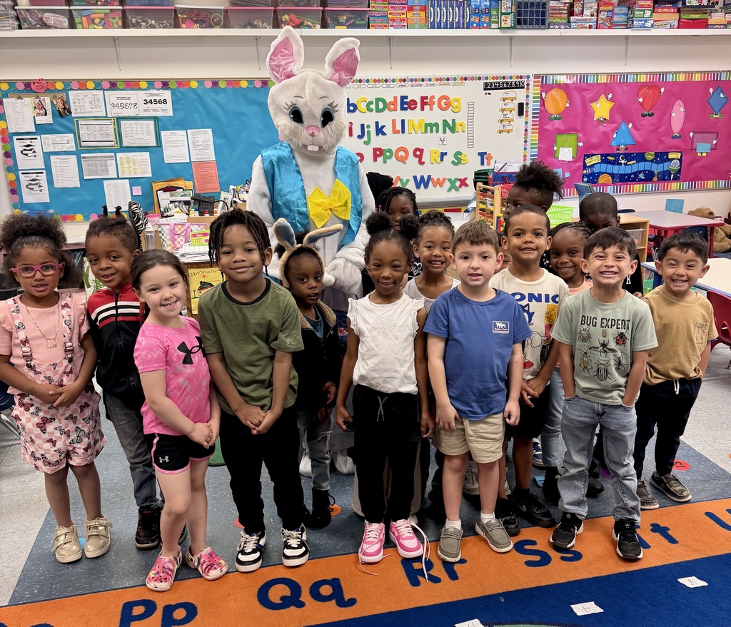 Students in a classroom  standing around the Easter Bunny, who is wearing a classic bunny costume. The children are smiling.