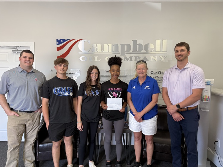 Six people stand together in an office in front of a “Campbell Oil Company” sign, with one student holding a donation check while others smile beside her.
