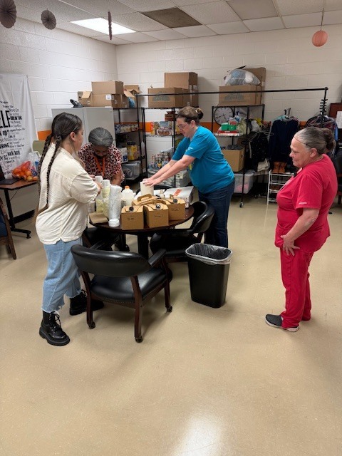 Four women stand around a table in a school resource room, organizing and distributing food items and supplies. Shelves in the background are stocked with boxes, canned goods, and clothing, indicating a pantry or support space for students and families.