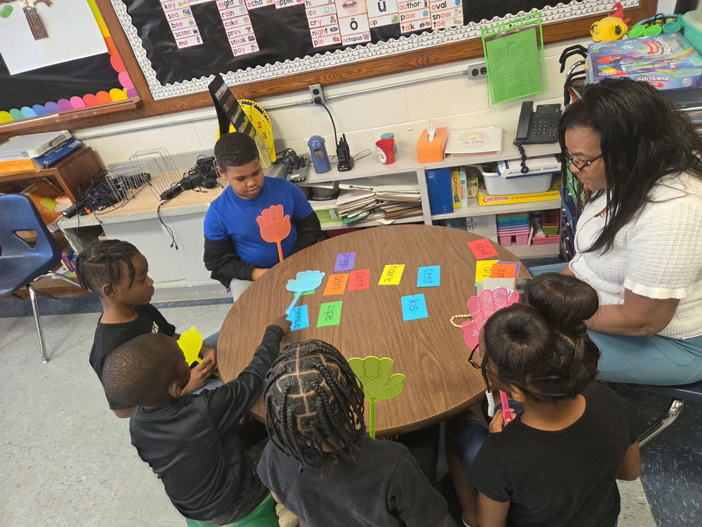 A teacher works with first grade students in a small group phonics lesson as students identify spelling words placed on the table during an interactive word recognition game.