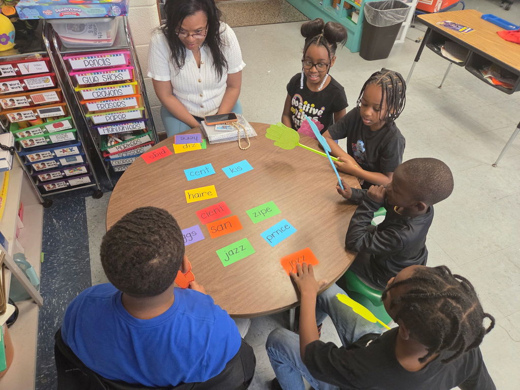 Students participate in a hands-on spelling activity at a small group table, using fly swatters to select spelling words from colorful cards as their teacher guides instruction.