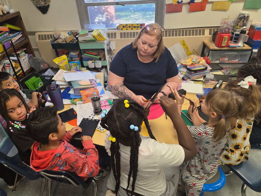 First grade students gather around a teacher at a kidney table using writing tablets to practice spelling and phonics skills during small group instruction.