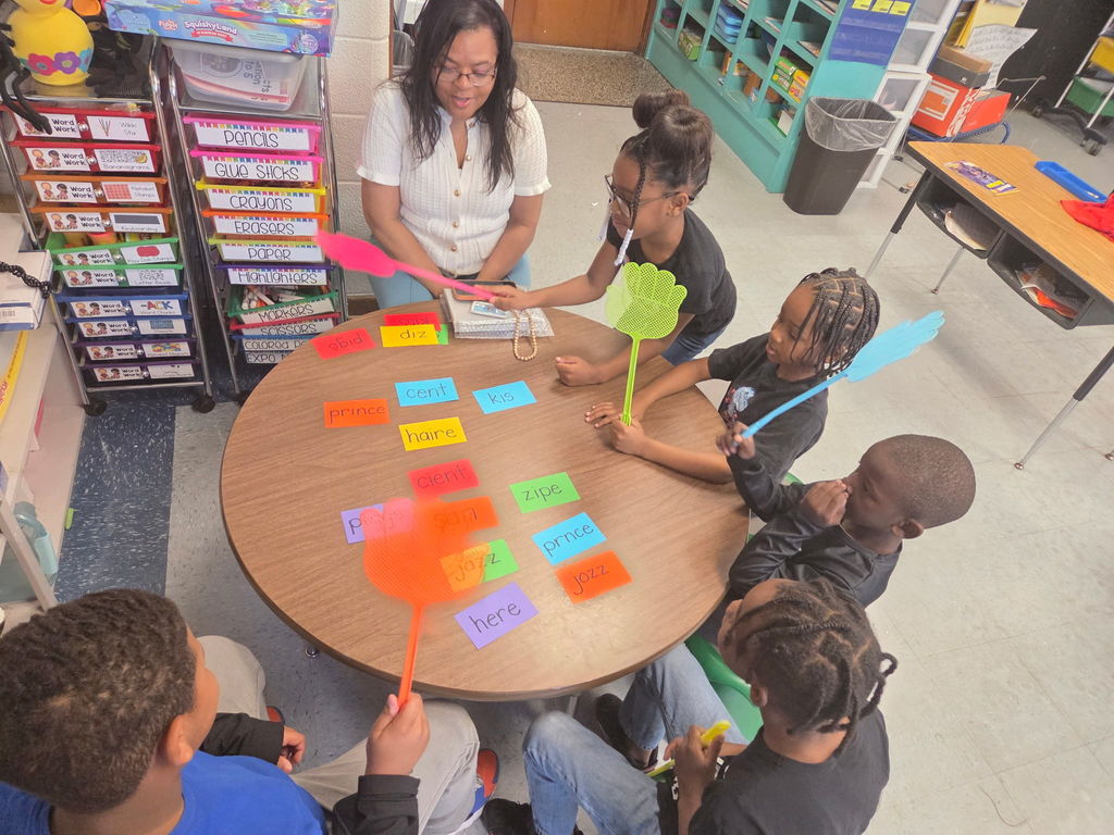 First grade students sit around a small group table practicing spelling words with colorful word cards while holding fly swatters to identify answers during a phonics game with their teacher.