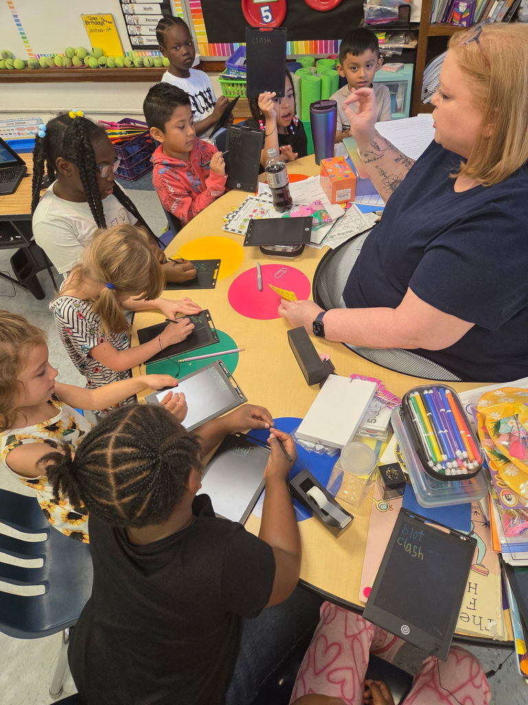 Students write spelling words on individual writing tablets while participating in guided phonics instruction with their teacher during first grade small group literacy practice.