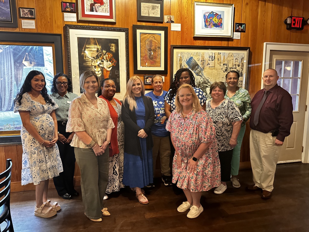 A group of eleven educators pose together and smile for a photo inside a restaurant with wood-paneled walls lined with artwork.