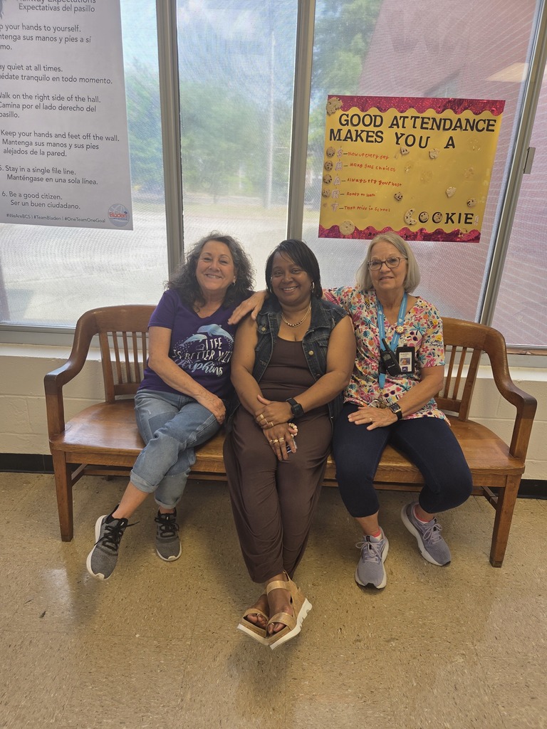 Three staff members sitting closely together on a wooden bench, smiling and showing friendship and support, with school posters visible in the background.