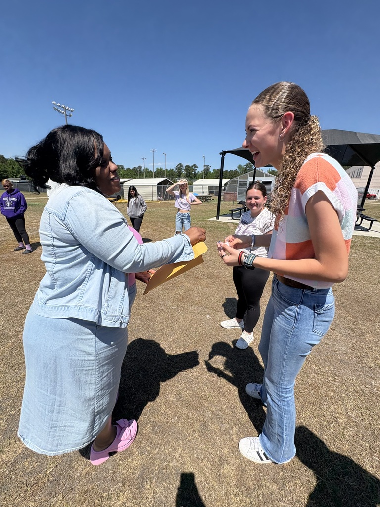 Honors English students stand in a circle on a grassy field during a Truth or Dare activity for Their Eyes Were Watching God. A school building and athletic lights are in the background.