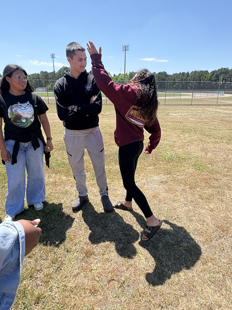Three students participating in an outdoor activity on a school field. One student in a maroon sweatshirt gives a high five to a classmate in a black hoodie.