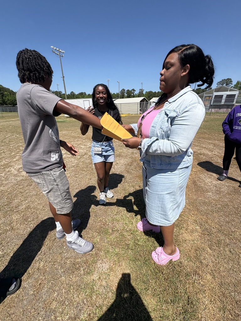 A student & teacher exchange a yellow envelope during an outdoor Honors English III "Truth or Dare" activity while a classmate smiles in the background