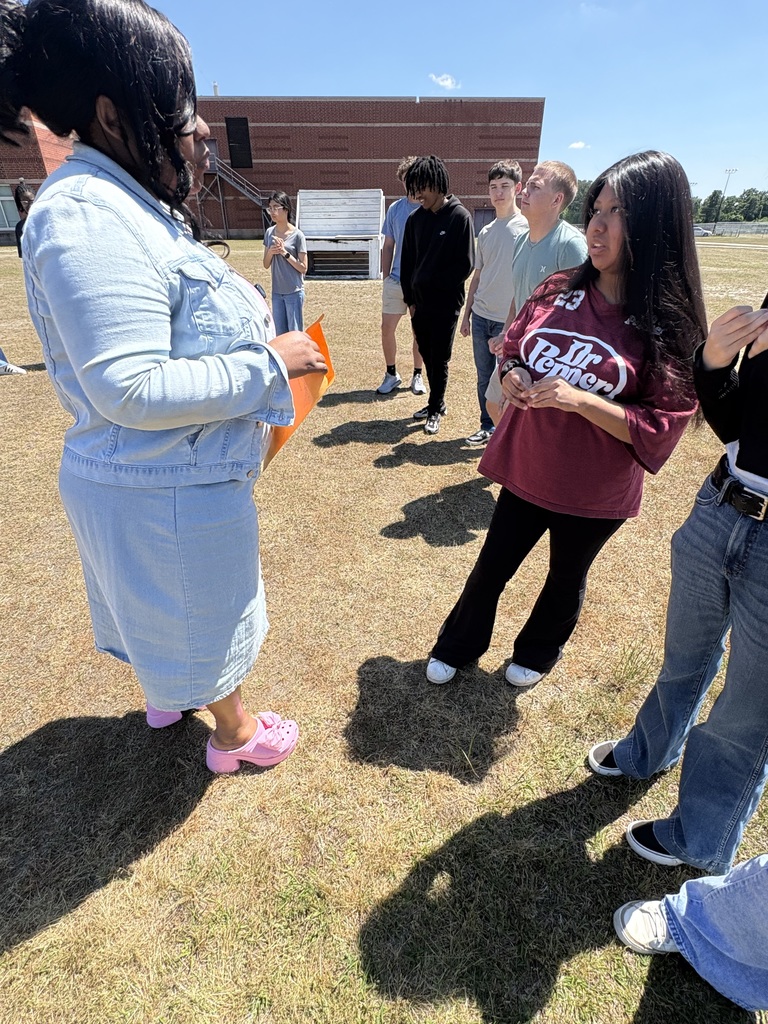 A group of high school students stands outdoors on a grassy area during a sunny day. They are engaged in a learning activity, with one student in a maroon Dr Pepper shirt talking to a teacher.