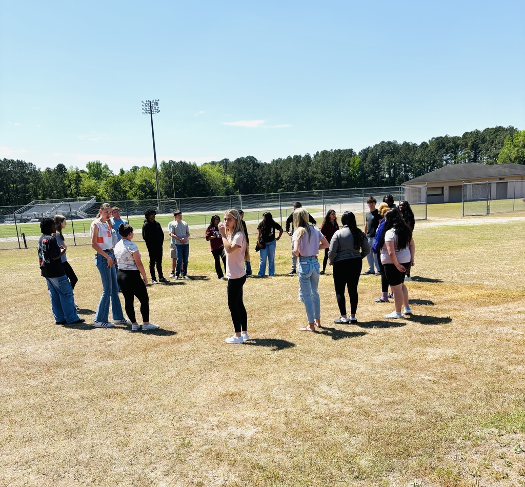 Honors English students stand in a circle on a grassy field during a Truth or Dare activity for Their Eyes Were Watching God. A school building and athletic lights are in the background.