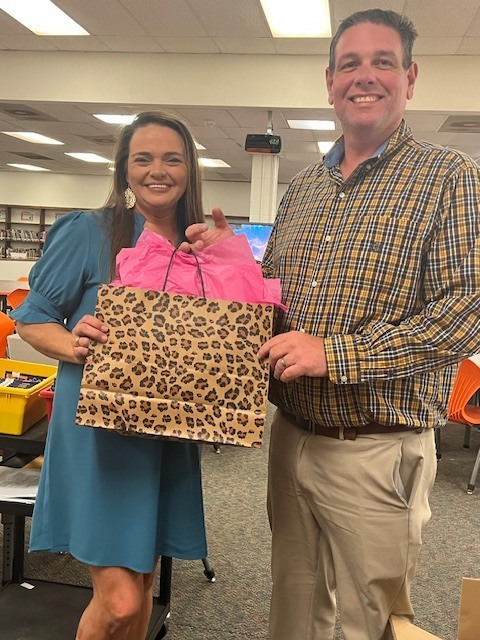Two adults stand in a school classroom smiling while holding a gift bag with a leopard print design and pink tissue paper. Tables, chairs, and classroom materials are visible in the background.