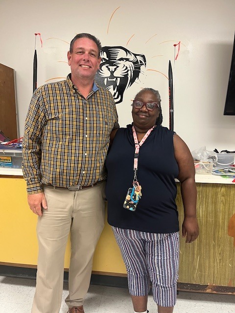 wo staff members stand together smiling in a school classroom. Behind them is a wall with a cougar mascot graphic, and classroom materials and supplies are visible on a counter nearby.