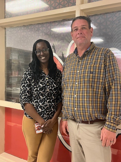 Two staff members stand side by side smiling in a school hallway. One is wearing a patterned blouse and glasses with an ID badge, and the other is wearing a plaid shirt and khaki pants. A wall with school colors and a cougar logo is visible behind them.