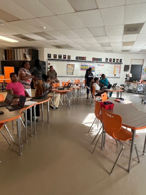 Participants gather and discuss while using laptops in a classroom, working together in small groups.