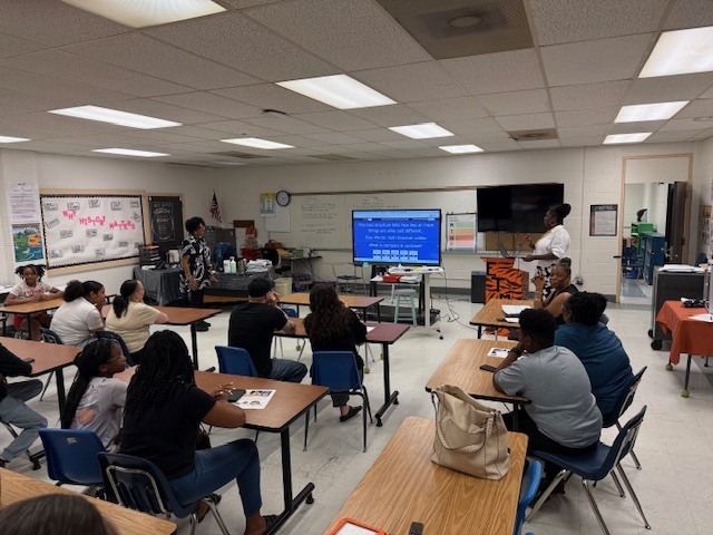 Another view of the classroom shows participants listening and interacting as a presentation is displayed at the front of the room.