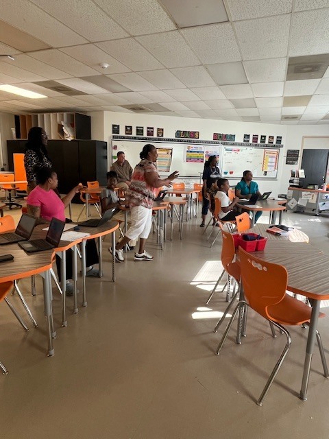Staff members stand and collaborate around tables with laptops in a classroom with orange chairs during a hands-on session.